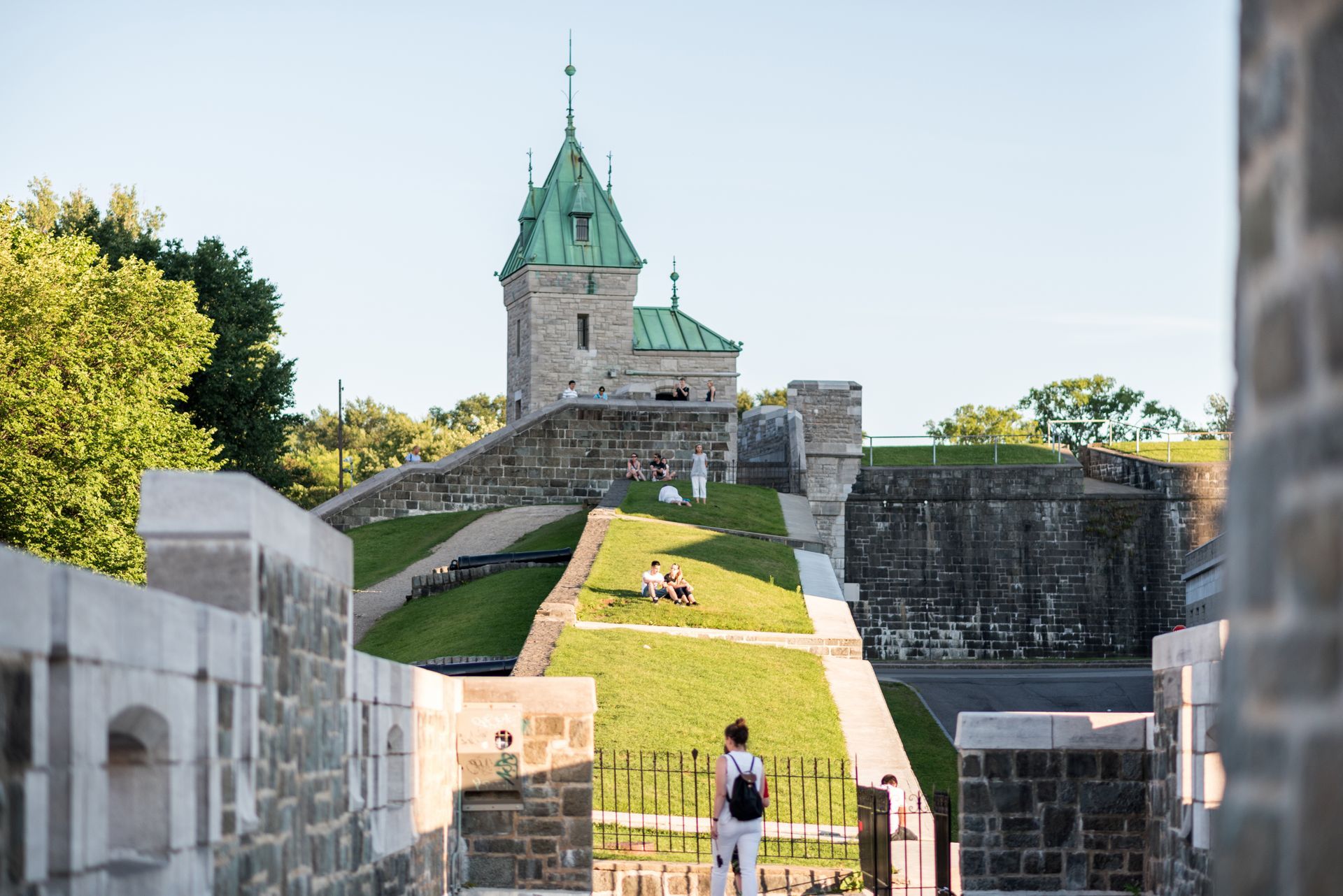 De stadsmuren van Old Quebec met een toren met een groen dak en een met gras begroeide wal, met een persoon die eronder loopt.