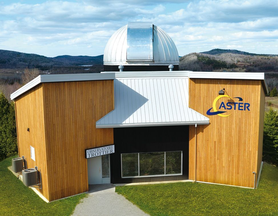 Het observatoriumgebouw ASTER in Quebec, Canada, met een zilverkleurige koepel en een met hout beklede buitenkant, gelegen op een met gras begroeide heuvel.