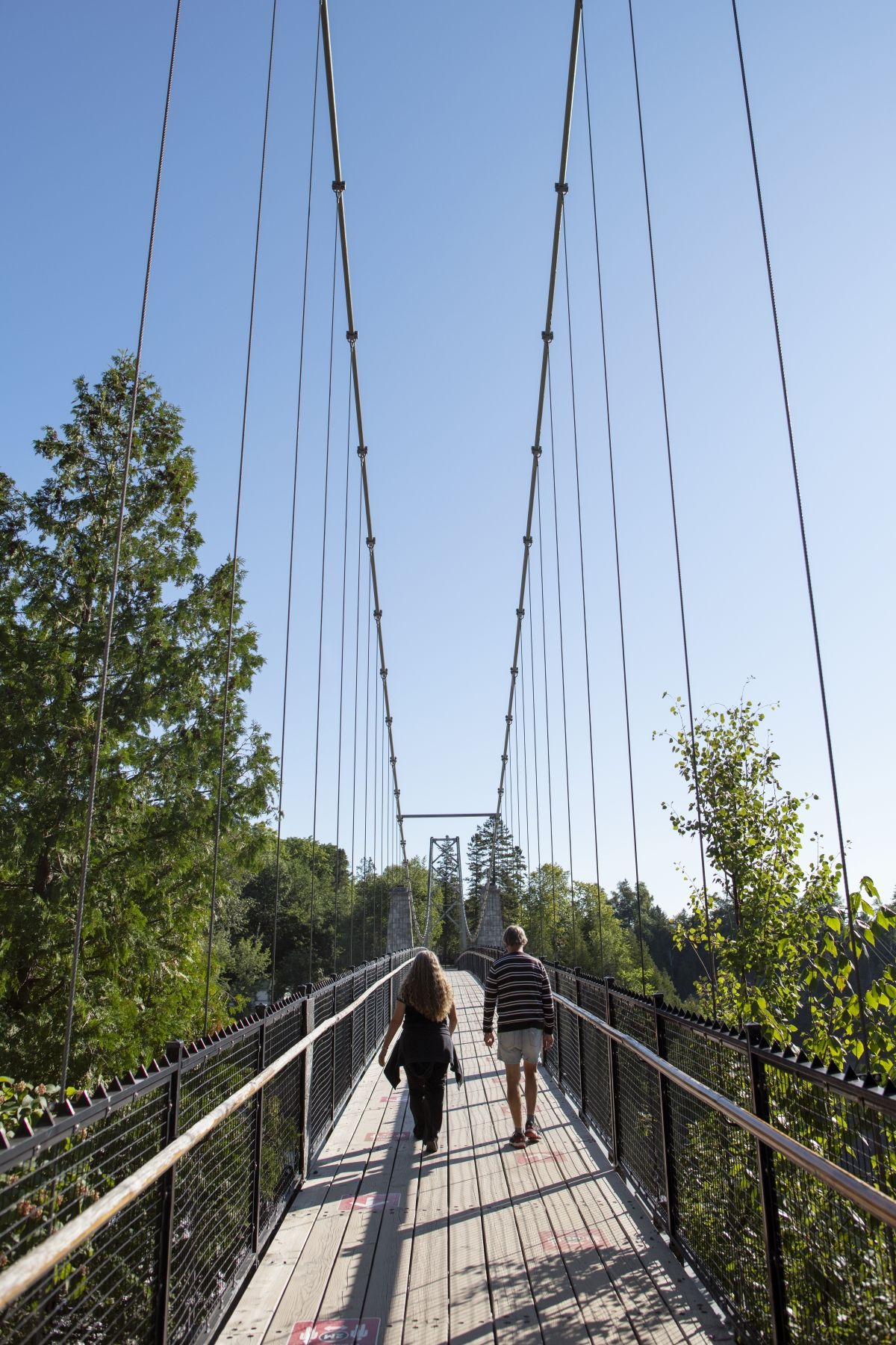Een hangbrug voor voetgangers in Parc de la Chute-Montmorency in Canada met twee mensen die onder een blauwe hemel lopen, omringd door bomen.