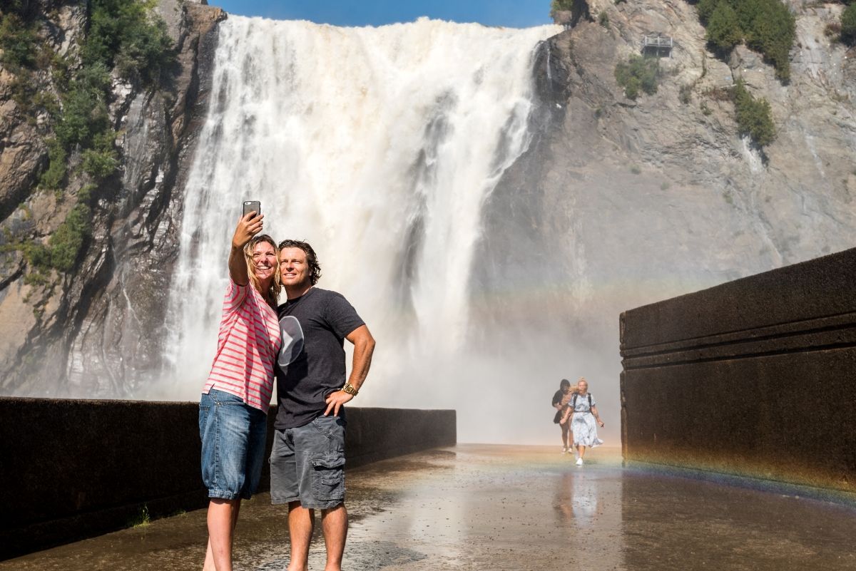 Een stel poseert voor de grote waterval in Parc de la Chute-Montmorency in Canada met mist en een regenboog.