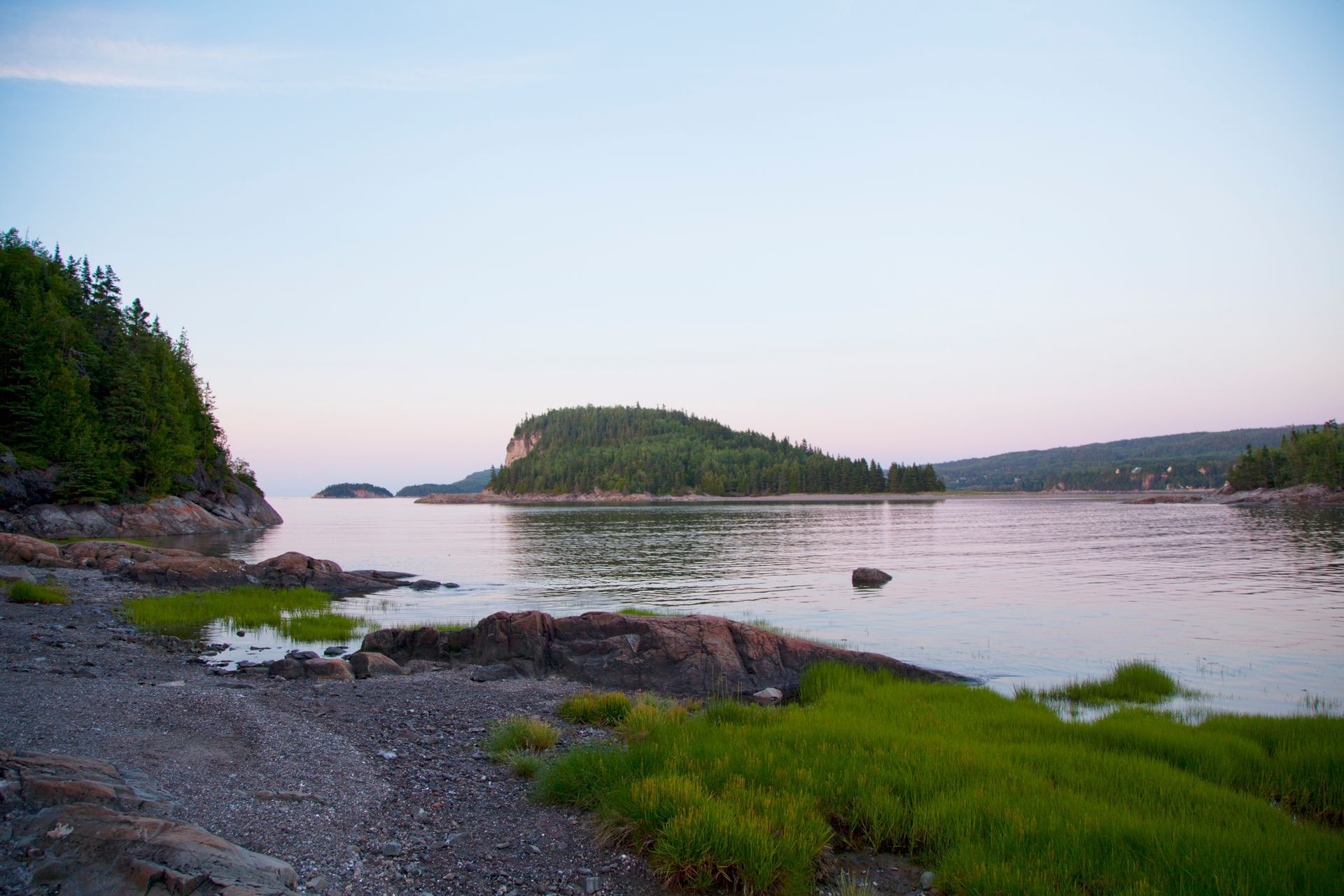 De rotsachtige kustlijn van in Parc national du Bic in Quebec, Canada, met groen gras, kalm water en een klein, met bomen begroeid eilandje bij zonsondergang.