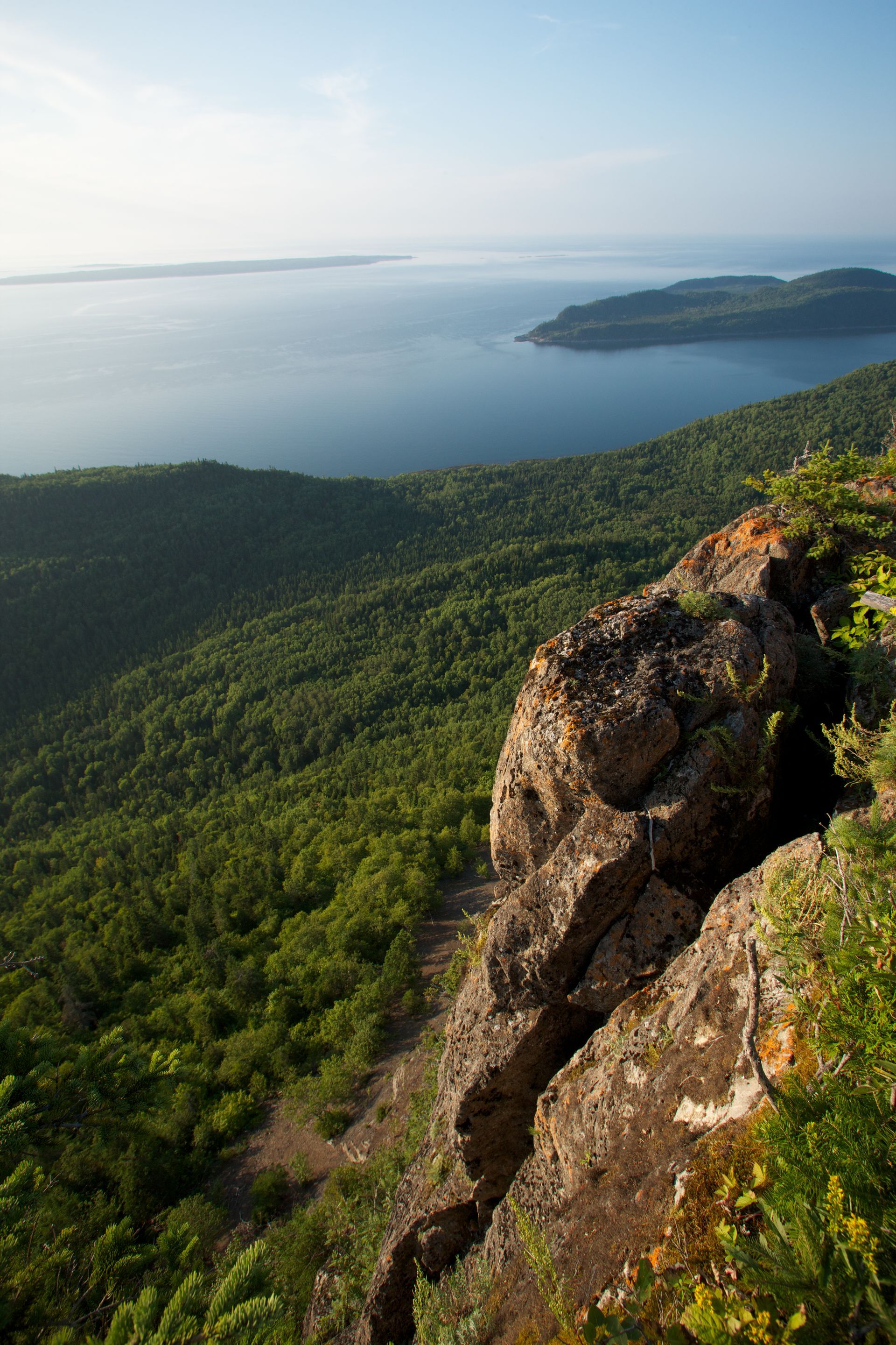 Zonnig uitzichtpunt aan de kust in Parc national du Bic, Quebec Canada, boven een beboste heuvel en een kalme blauwe zee, met een rotsachtige klif op de voorgrond.