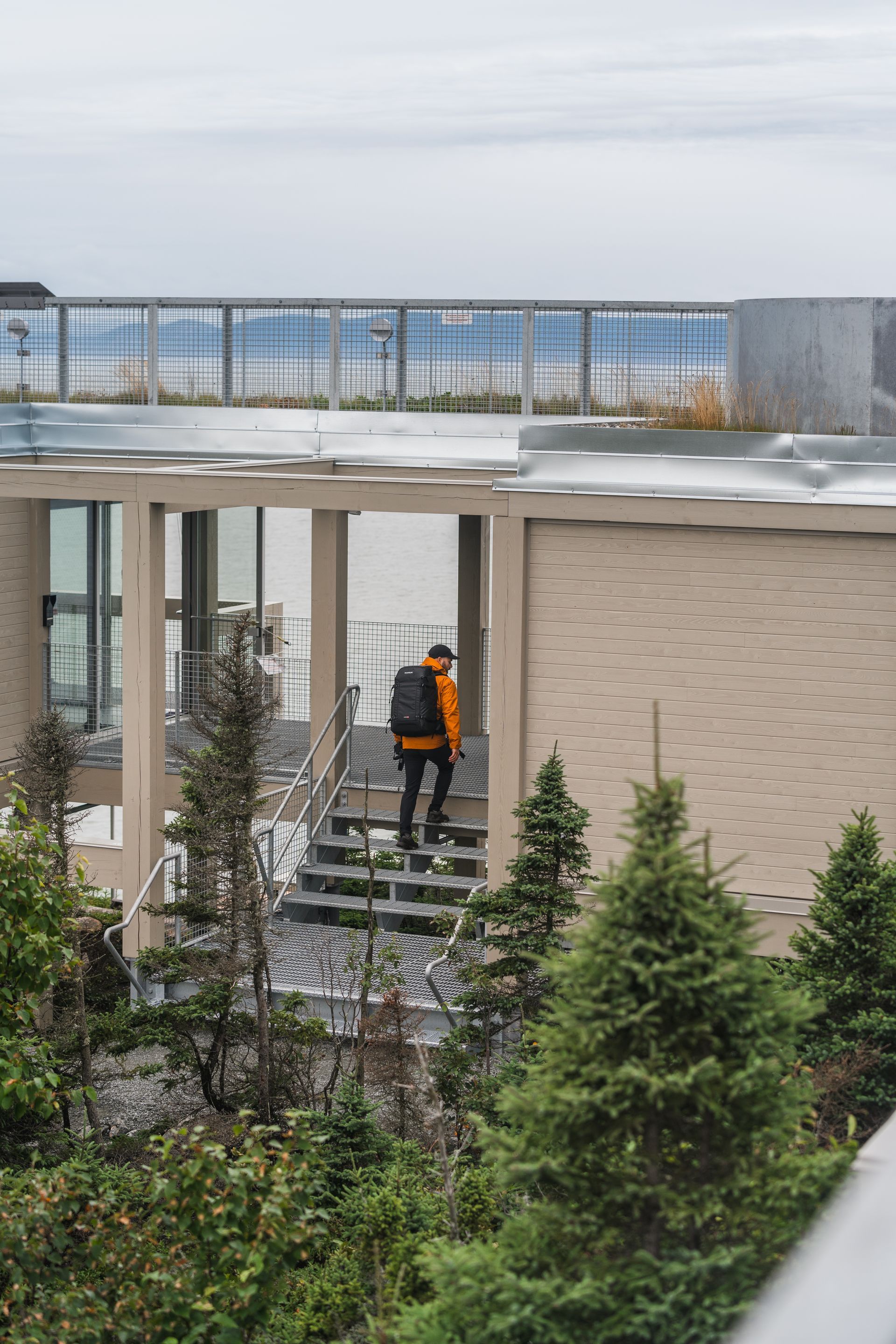 Een persoon in een oranje vest beklimt een trap naast Putep ’t-awt Cacouna observation site in Quebec, Canada, met groenblijvende bomen ervoor.