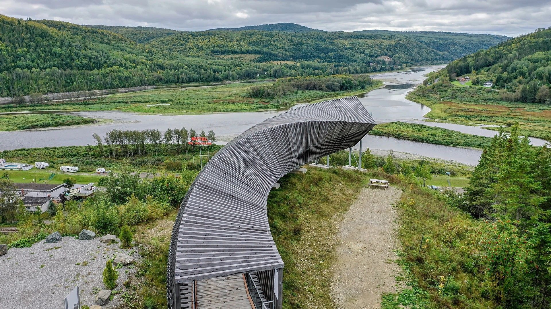 Een houten vlonderpad slingert door een groene vallei langs een rivier en beboste heuvels.