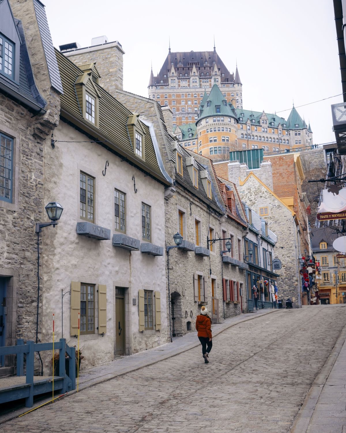 Een smalle straat in Québec, Canada, met kinderkopjes, geflankeerd door oude stenen gebouwen, met een persoon die richting een kasteel op een heuvel loopt.