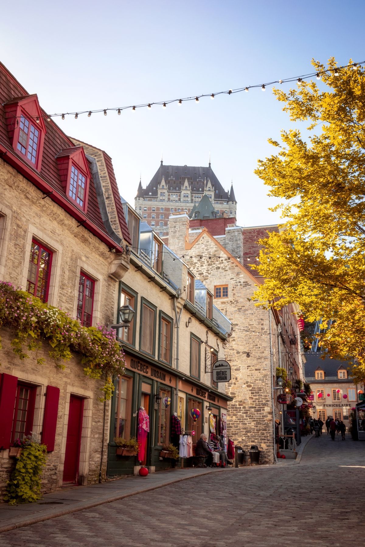 Een charmante straat in Québec, Canada, met kinderkopjes, kleurrijke oude gebouwen, herfstbomen en een kasteel op een heuveltop in de verte onder een blauwe hemel.