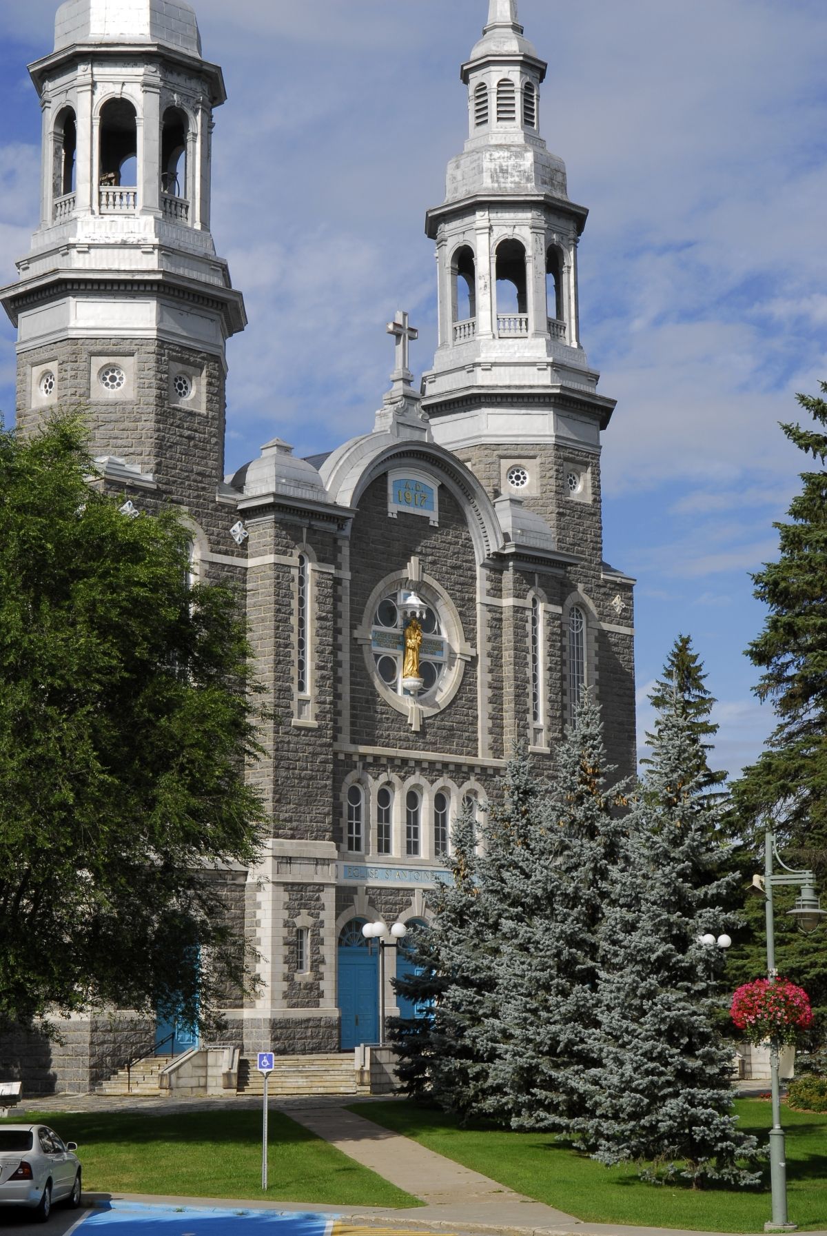 De Saint-Antoine-de-Padoue kerk in
Louiseville in Canada met twee torens en een cirkelvormige muurschildering, omgeven door bomen en een gazon onder een blauwe hemel.