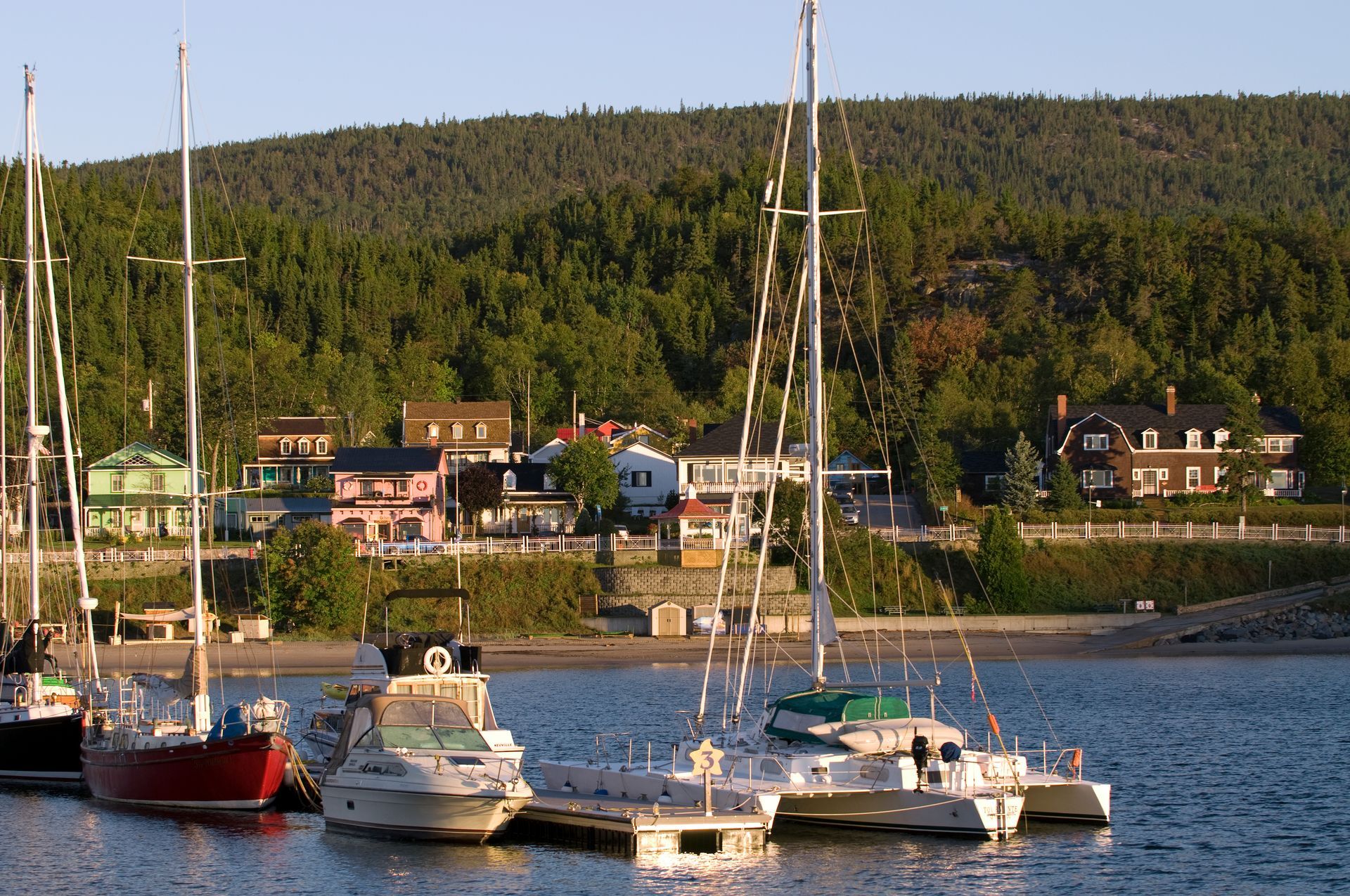 Verschillende zeilboten liggen aangemeerd in de haven van Tadoussac in Canada met huizen aan het water en een beboste heuvel op de achtergrond.