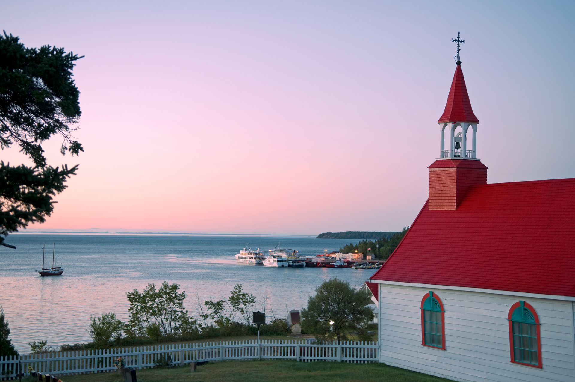 Kerk met rood dak van Tadoussac in Canada aan een rustige baai bij zonsondergang, met pastelkleurige lucht en kustlijn in de verte.