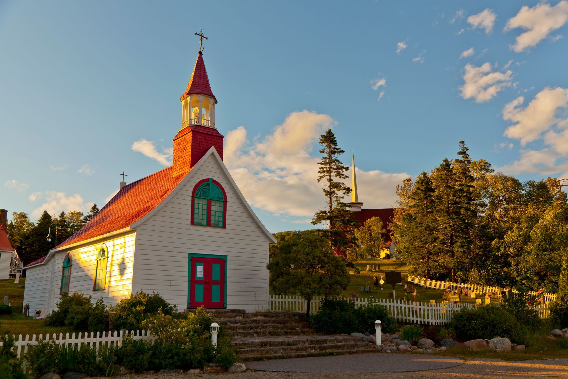 Wit kerkje in Tadoussac in Canada met rood dak en torenspits in een tuin bij zonsondergang, omgeven door bomen en een wit hek.