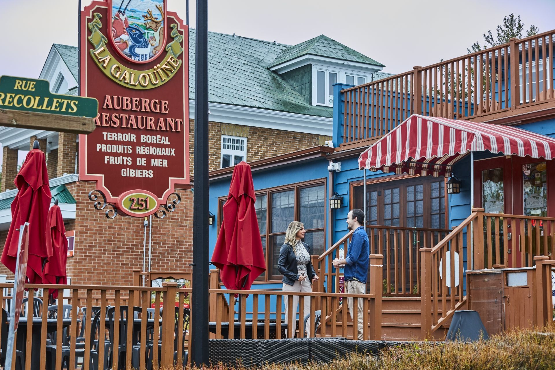 Bakstenen restaurant in Tadoussac, Canada, met groen dak en gestreepte luifel, rode parasols en twee mensen op het terras.