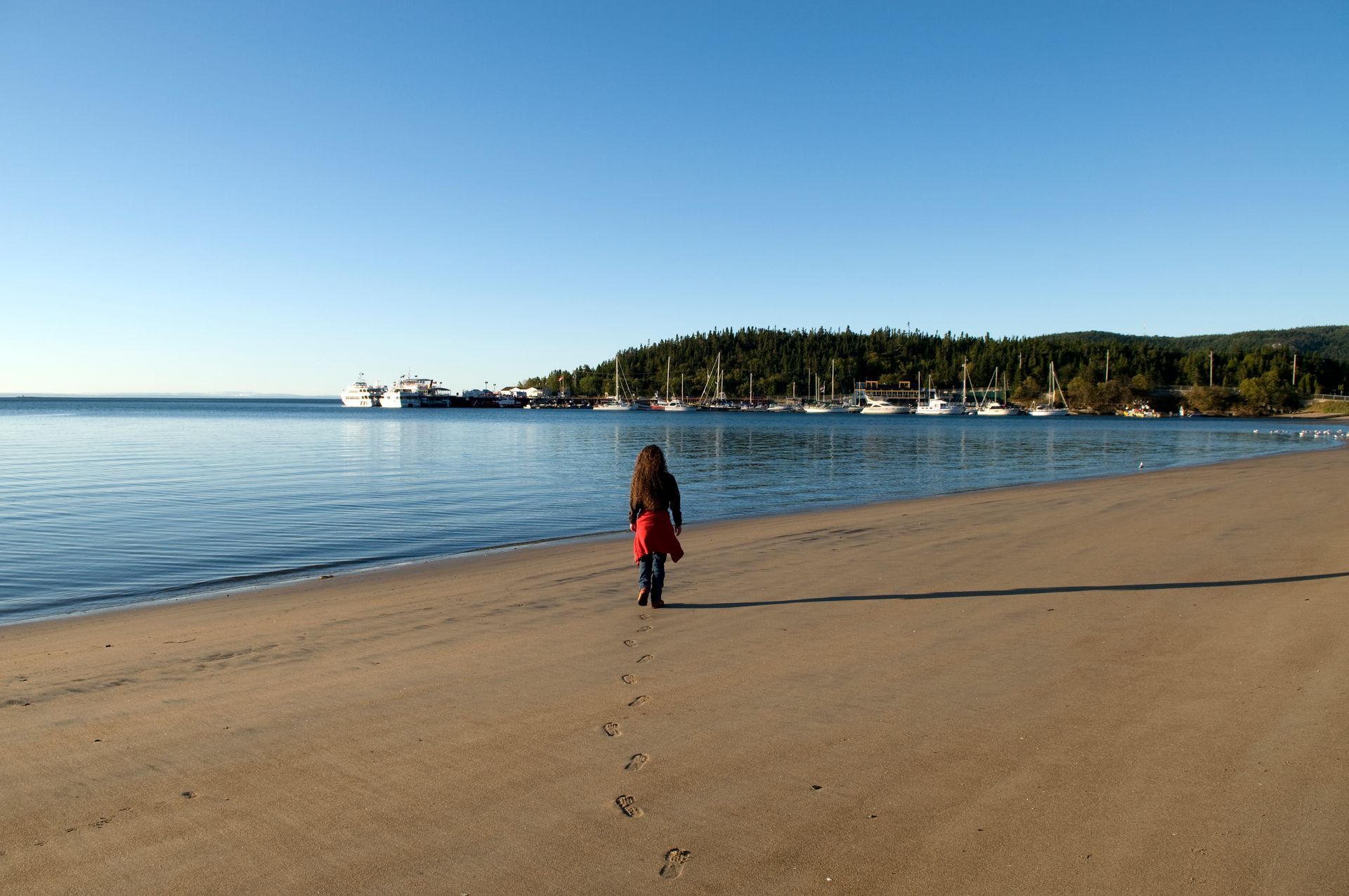 Een persoon in een rode jas loopt over het zandstrand van Tadoussac in Canada langs kalm water op een heldere dag.