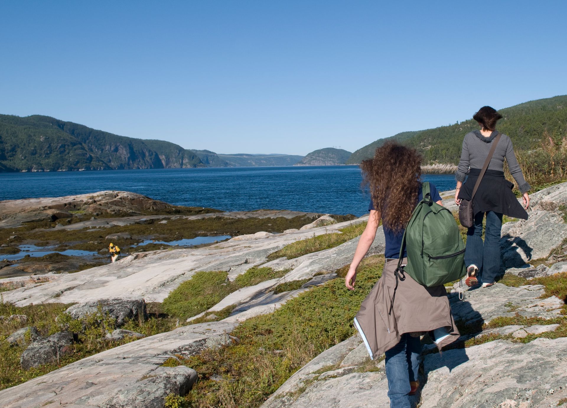 Twee wandelaars lopen langs een rotsachtige kustlijn in Tadoussac, Canada, met blauw water en groene heuvels op de achtergrond.