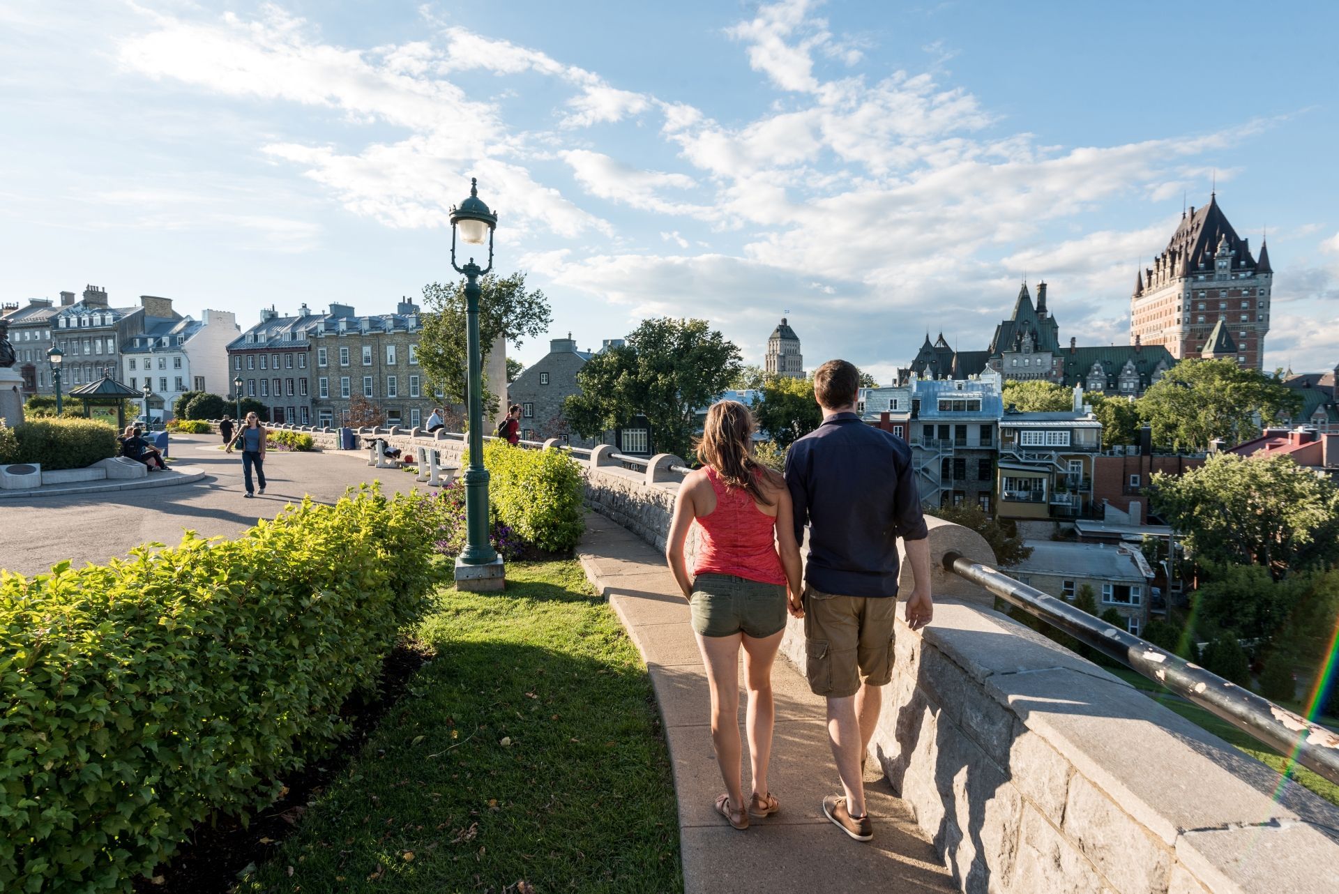 Twee mensen lopen over een pad op een heuveltop langs een rivier, met in de verte een stadsgezicht en een kasteel in Québec.