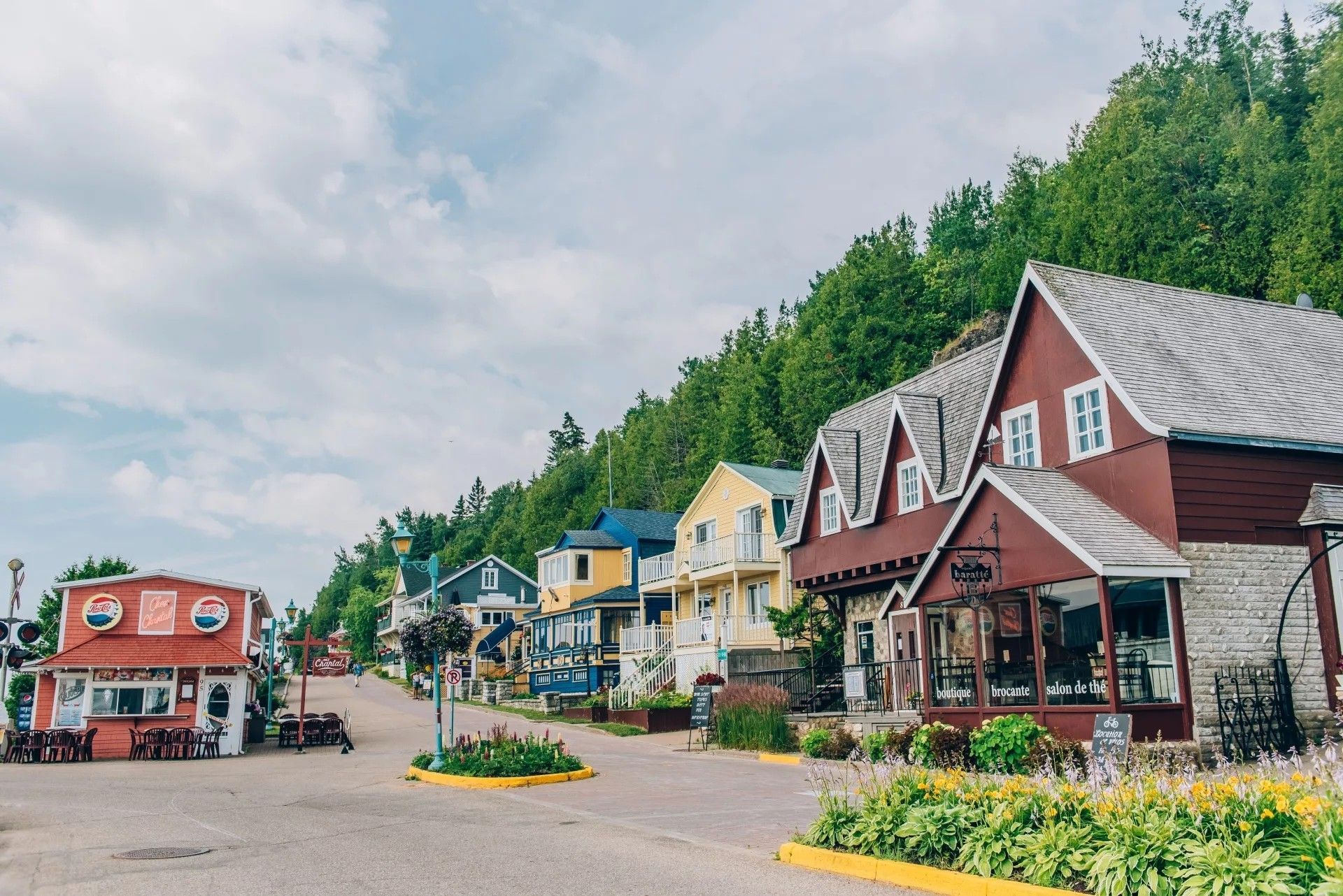 Een kleurrijke dorpsstraat in La Malbaie, Canada, met schilderachtige huisjes, bloemperken en een beboste heuvel onder een bewolkte hemel.