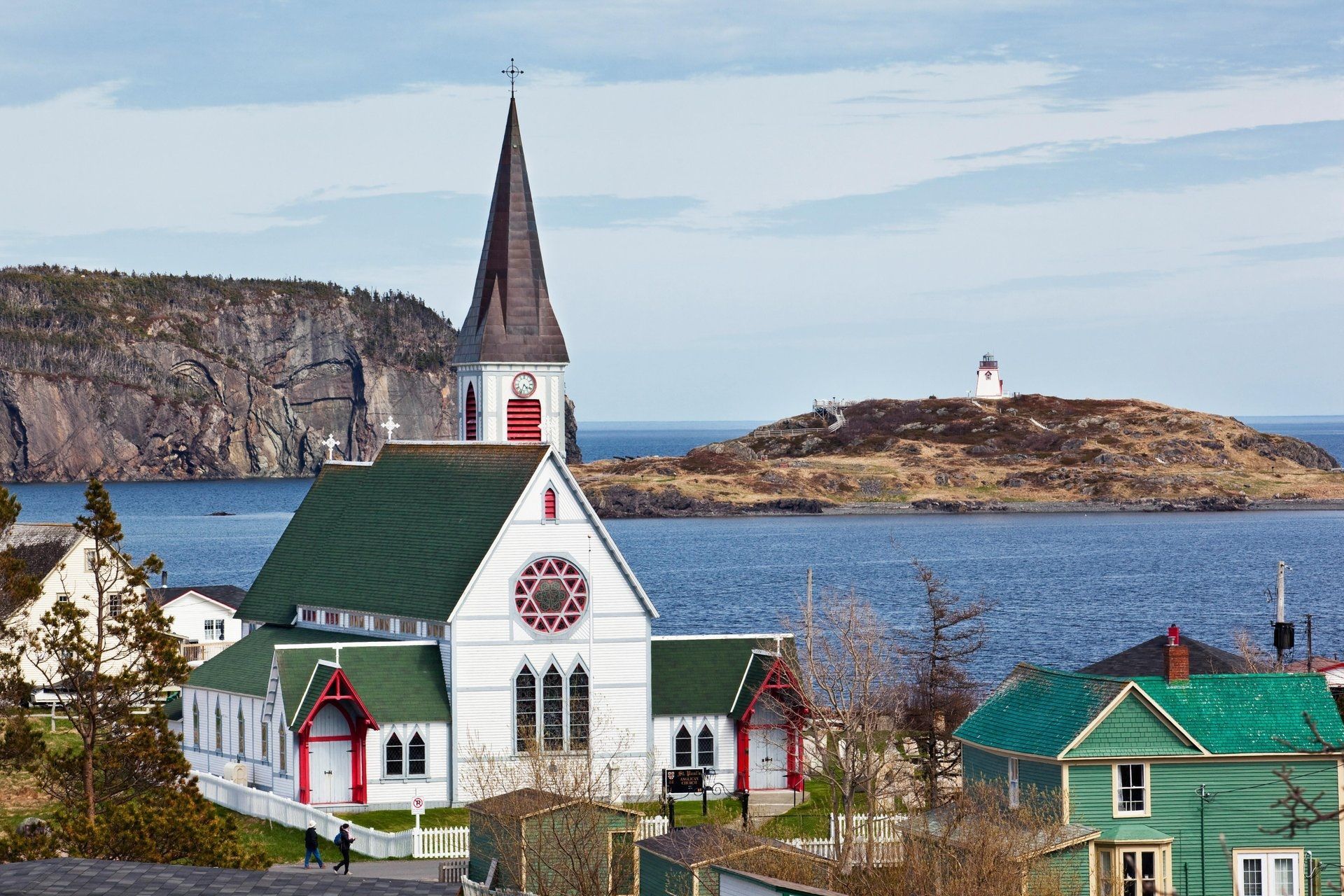Witte kerk in Newfoundland, Canada met groene daken aan zee, met een rotsachtig eiland en een vuurtoren op de achtergrond.