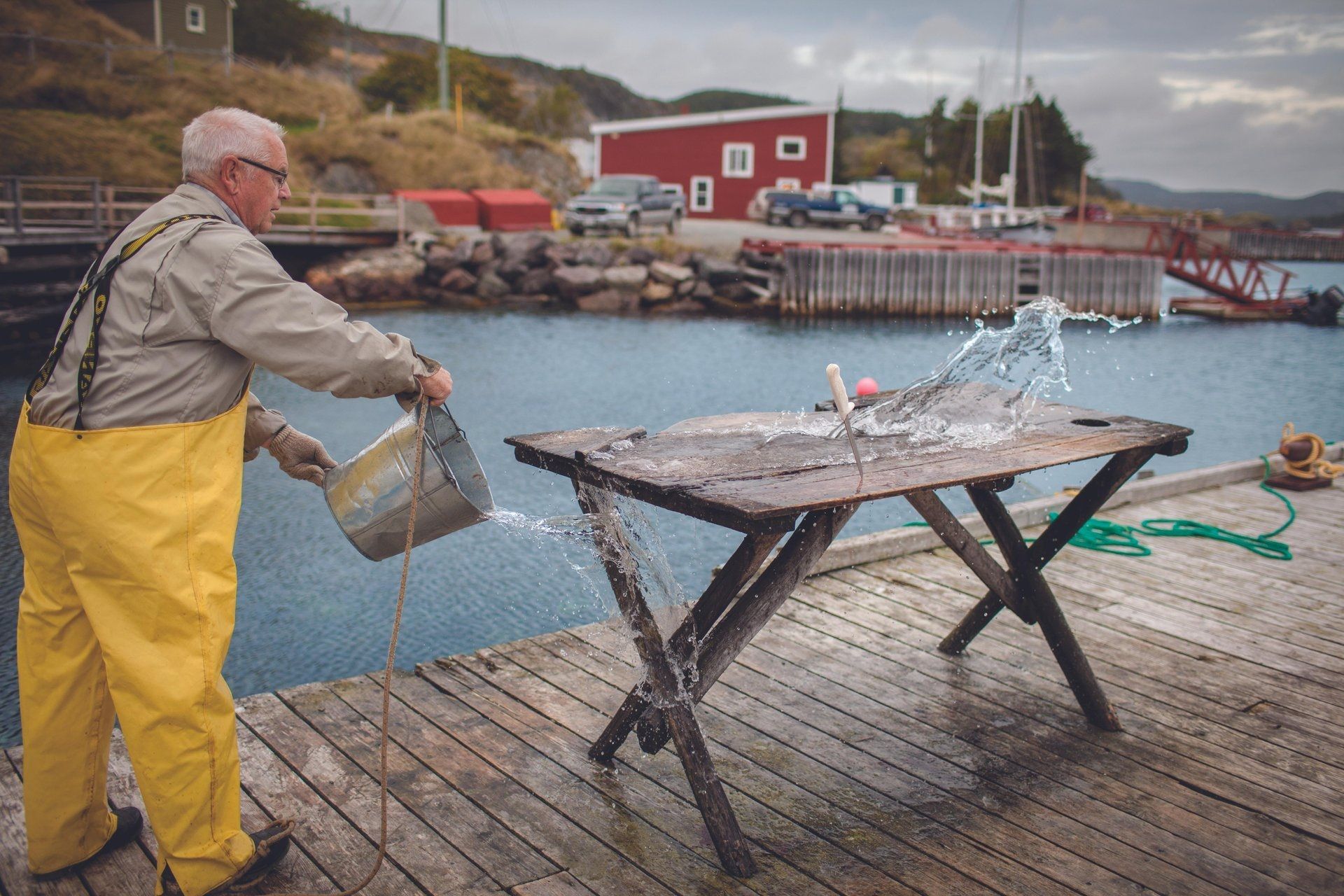Een visser in Newfoundland Canada in een gele overall spoelt een vis af op een kade naast een haven met rode gebouwen.