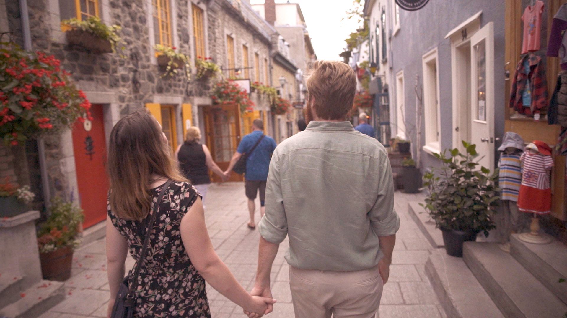 Een stel loopt hand in hand door een smalle, met bloemen omzoomde straat met winkels en kleurrijke gebouwen in Old Quebec.