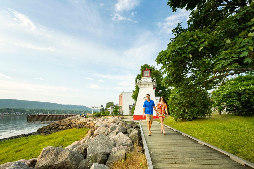 Twee mensen lopen in Annapolis Royal , Nova Scotia, over een houten vlonder langs de zee, langs rotsen en een wit, tempelachtig bouwwerk.