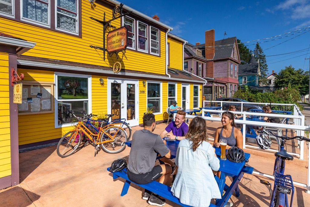 Mensen zitten aan een blauwe picknicktafel buiten bij felgele bakkerij van Annapolis Royal met geparkeerde fietsen.