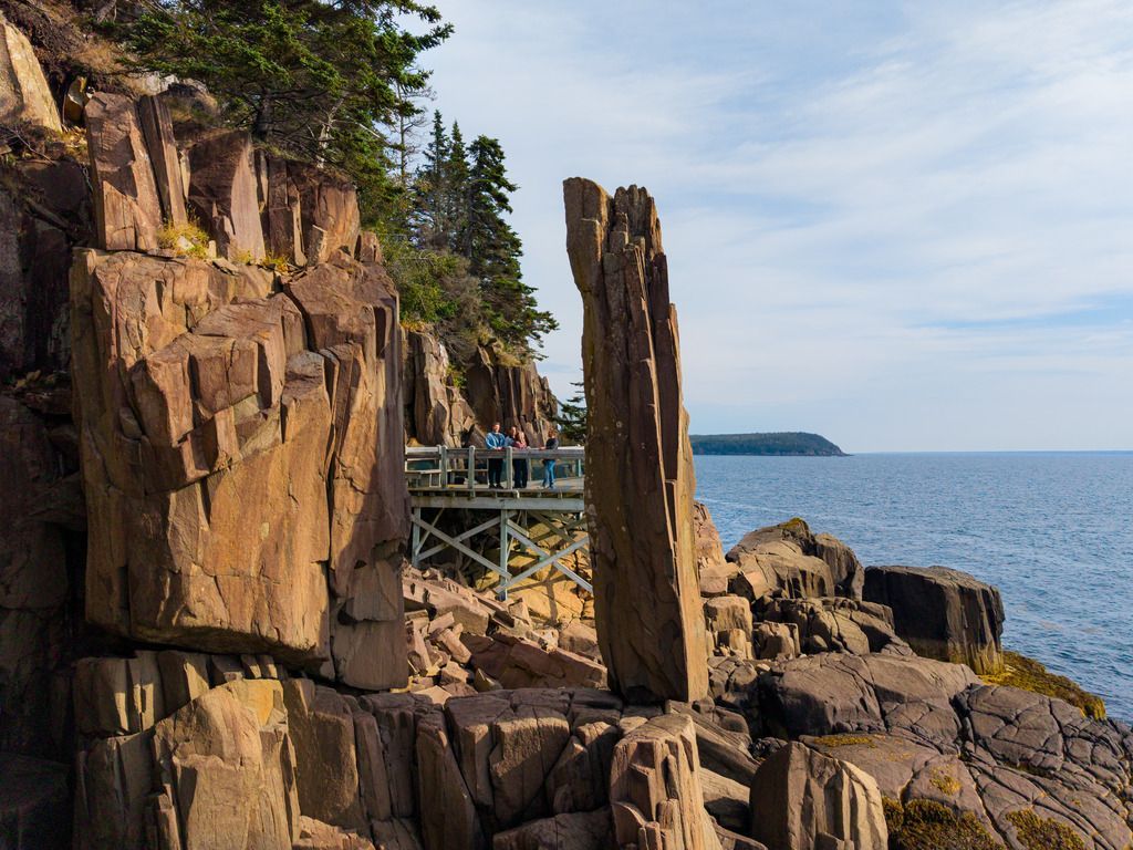 Een rotsachtige klif met Balancing Rock op Nova Scotia, gelegen naast kalm blauw water onder een gedeeltelijk bewolkte hemel.