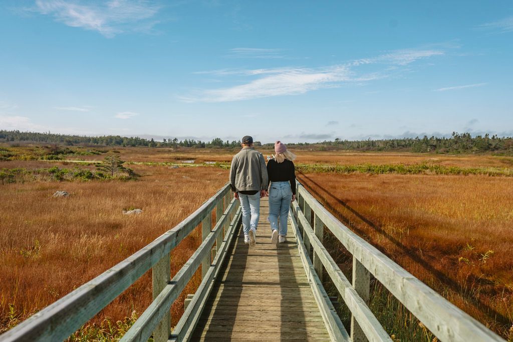 Twee mensen lopen over een houten vlonderpad op de Big Meadows Bog Trail op Brier Island, Nova Scotia, door een moeras onder een blauwe hemel.