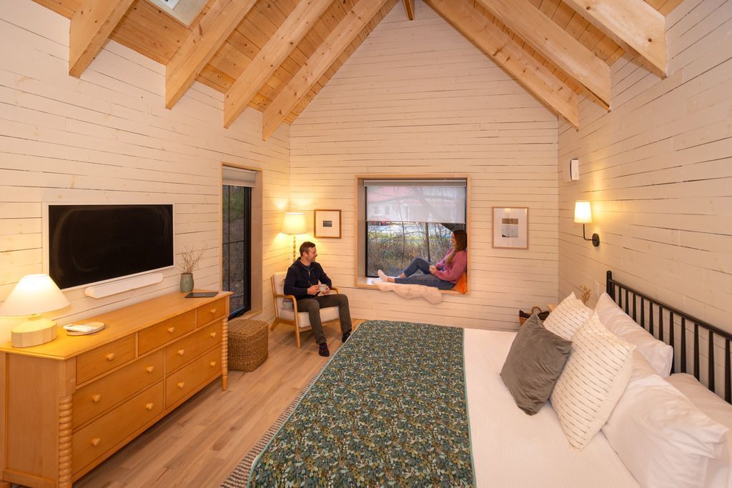 Gezellige loftslaapkamer van Black Forest Cabins in Mahone Bay op Nova Scotia met gewelfd houten plafond, bed, commode, tv en een persoon die bij het raam zit.