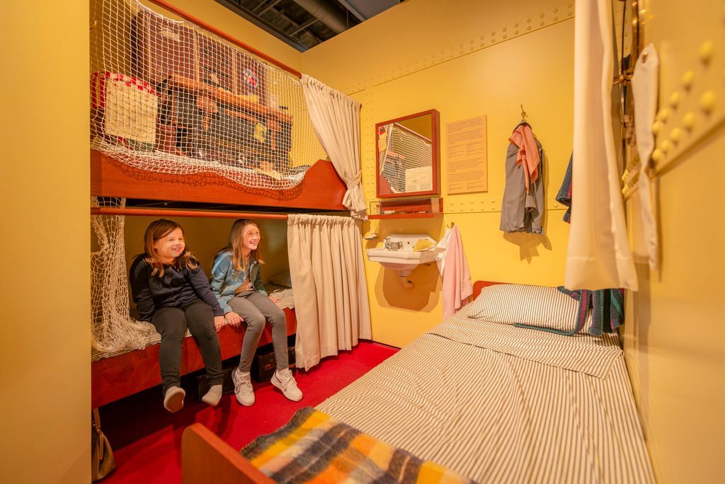 Twee kinderen zitten in het Canadian Museum of Immigration te Halifax op Nova Scotia, Canada, op een bed in een gezellige gele slaapkamer met een rood vloerkleed en gestreept beddengoed.
