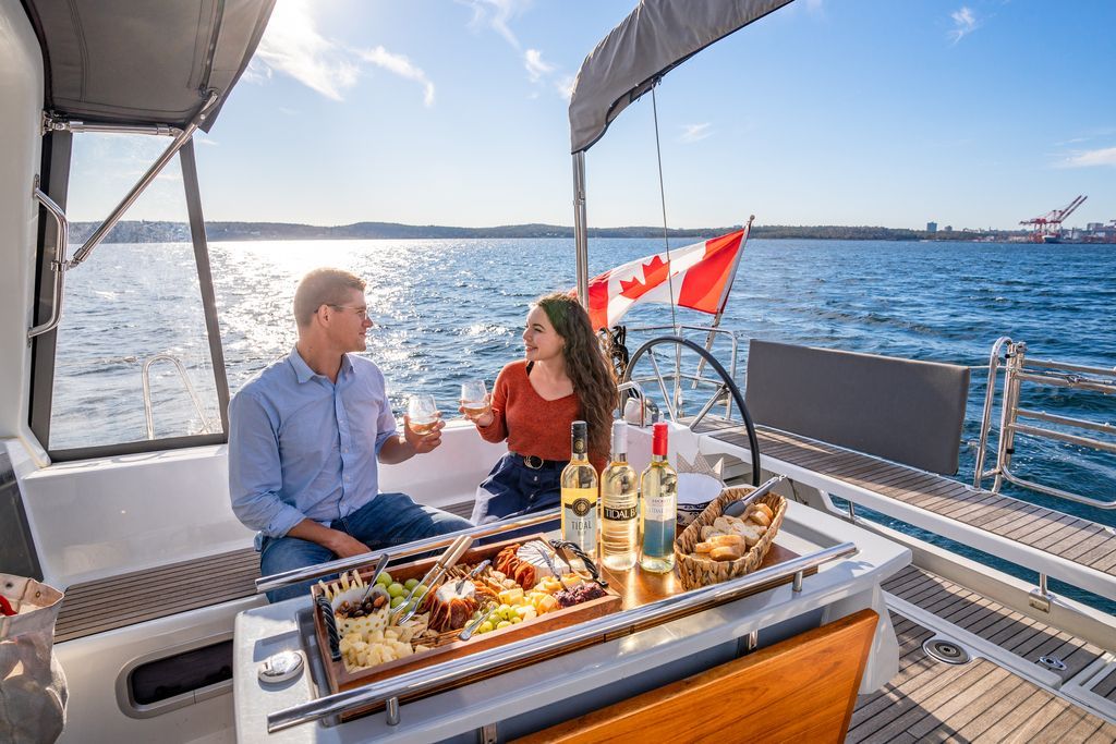 Twee mensen dineren op het dek van een boot met op de achtergrond de skyline van Halifax op Nova Scotia, Canada, en rode vlaggen.