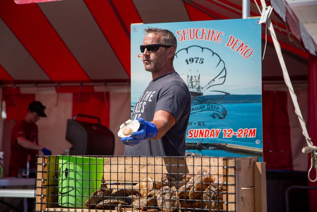 Een man bij een viskraam in Digby op Nova Scotia houdt Sint Jacobsschelpen vast naast een bord met reclame voor verse vis en openingstijden op zondag.