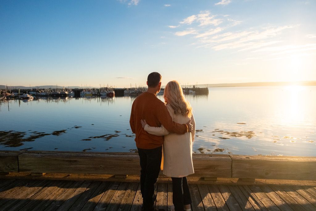 Een stel staat in Digby, Nova Scotia, op een steiger bij zonsondergang en kijkt uit over een kalm meer met heldere weerspiegelingen.