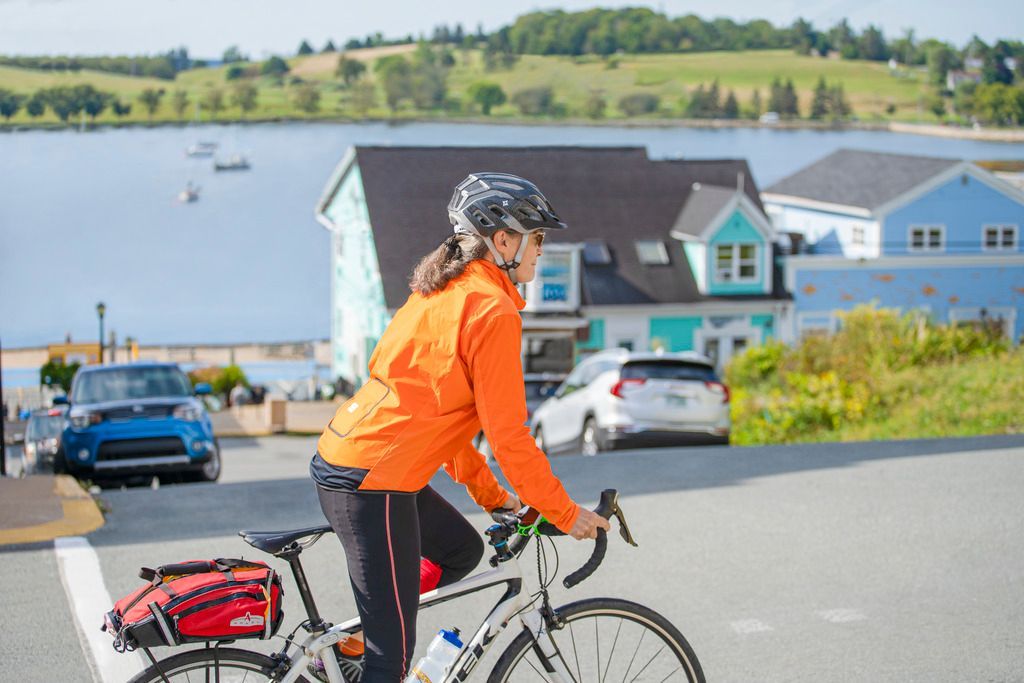 Een fietser in een oranje jas rijdt in Lunenburg, Nova Scotia, langs een weg aan het water, met huizen en geparkeerde auto's in de buurt.