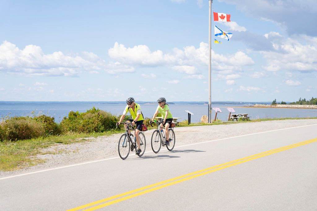 Twee fietsers in neonkleurige hesjes rijden langs de zee in de omgeving van Lunenburg, Nova Scotia, op een zonnige weg, met een Canadese vlag in de buurt.