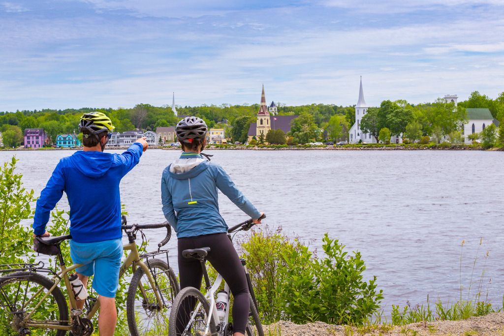 Twee fietsers in blauwe jassen pauzeren bij een meer en wijzen naar het stadje Mahone Bay, Nova Scotia, aan de overkant van het water.