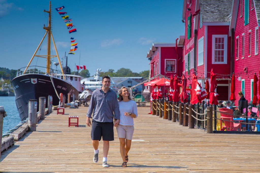 Een stel loopt over een zonnige houten promenade in Lunenburg, Nova Scotia, naast een aangemeerd schip en felroze gebouwen.