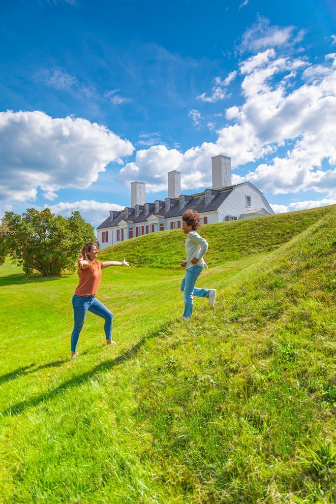 Twee kinderen spelen in het Port-Royal National Historic Site, Nova Scotia, op een grasheuvel onder een helderblauwe hemel, met een groot gebouw op de achtergrond.