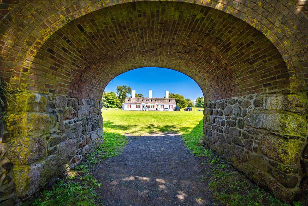 Uitzicht vanuit een stenen tunnel  in Fort Anne National Historic Site in Annapolis Royal, Nova Scotia, op een zonovergoten landhuis en een groen veld daarachter.