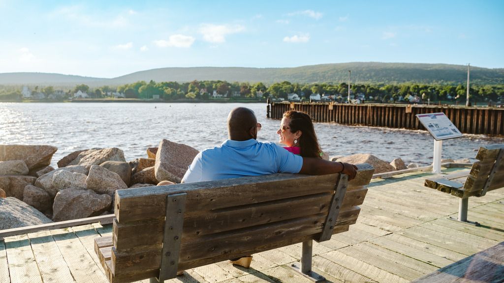 Een stel zit in Annapolis Royal, Nova Scotia, op een bankje aan een meer, met rotsen, een steiger en heuvels in de verte bij zonsondergang.