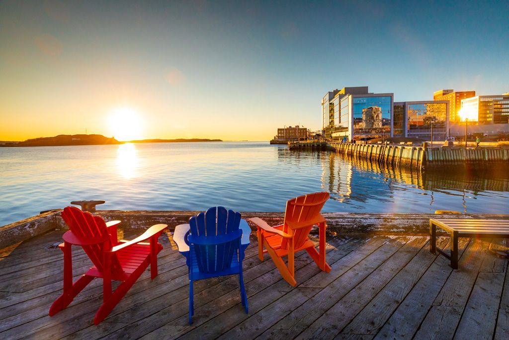 Kleurrijke Adirondack-stoelen op een steiger in Halifax op Nova Scotia in Canada bij zonsondergang, naast kalm water en gebouwen aan de waterkant.