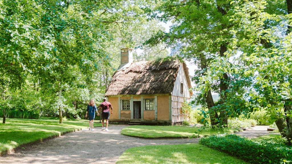 Klein huisje in de tuinen van de Annapolis Royal Historic Gardens in Annapolis Royal, Nova Scotia met twee mensen op een pad onder lommerrijke bomen.
