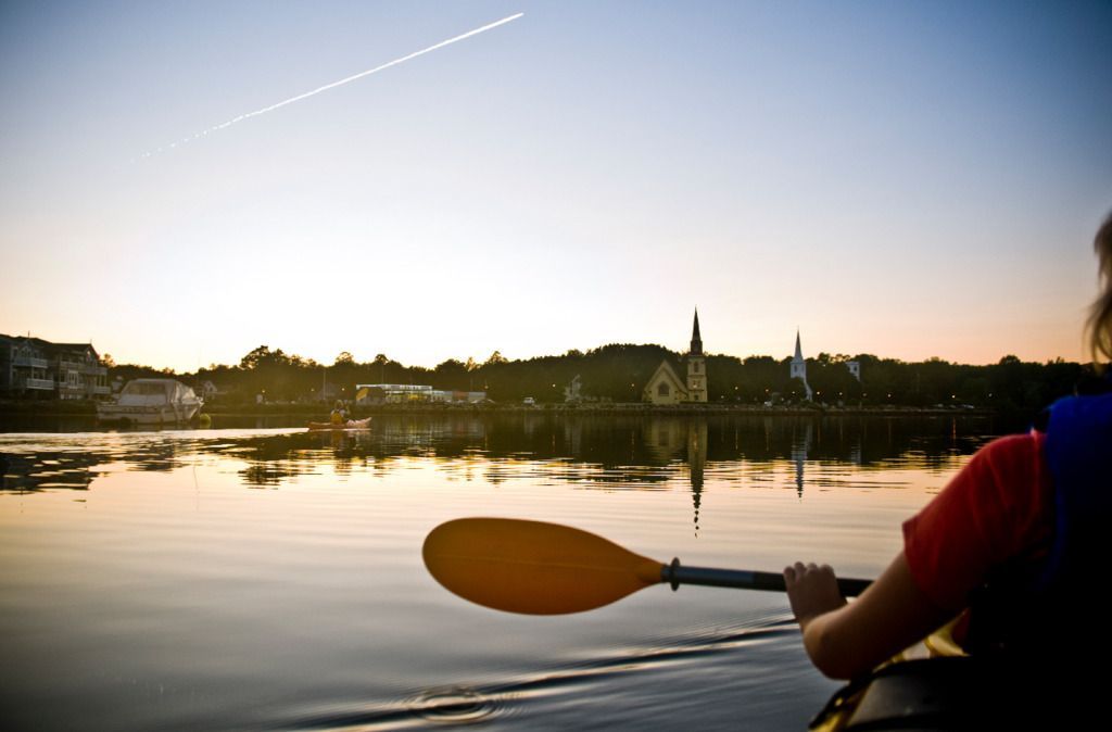 Een kajakker peddelt op een kalme zee bij Mahone Bay, Nova Scotia, bij zonsondergang, met een kerk en een rij bomen in de verte.