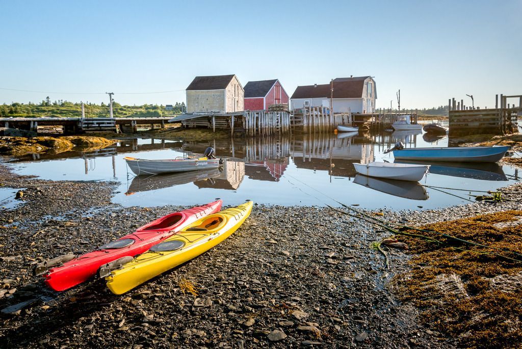 Twee kleurrijke kajaks op een kiezelstrand in Blue Rocks, Nova Scotai, naast een rustige haven met weerspiegelingen en huizen op de achtergrond.
