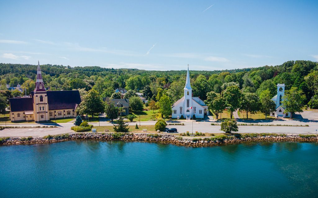 Het kleine dorpje Mahone Bay op Nova Scotia aan het water met een kerk, witte gebouwen en een blauw meer onder een heldere hemel.