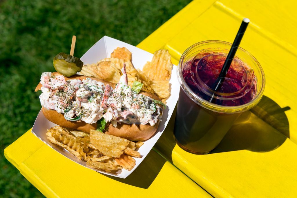 Een lobster roll en friet op een gele tafel in Lunenburg, Nova Scotia, naast een donker drankje met een rietje.