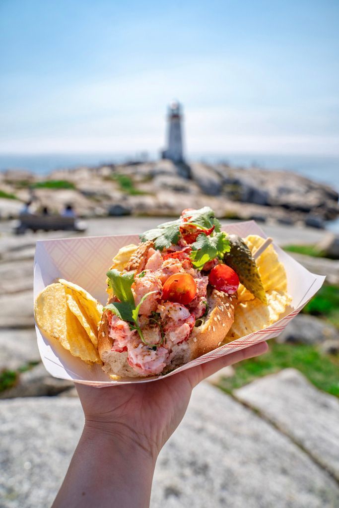 Een hand houdt een sandwich met zeevruchten en friet vast, met de vuurtoren van Peggy's Cove op Nova Scotia en een rotsachtige kustlijn wazig op de achtergrond.