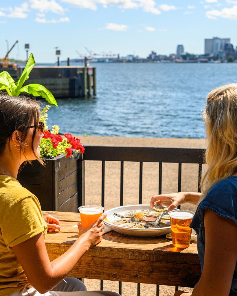 Twee mensen zitten gezellig te praten onder het genot van een drankje aan een tafeltje aan het water, met de skyline van de haven van  Halifax op Nova Scotia, Canada, op de achtergrond.