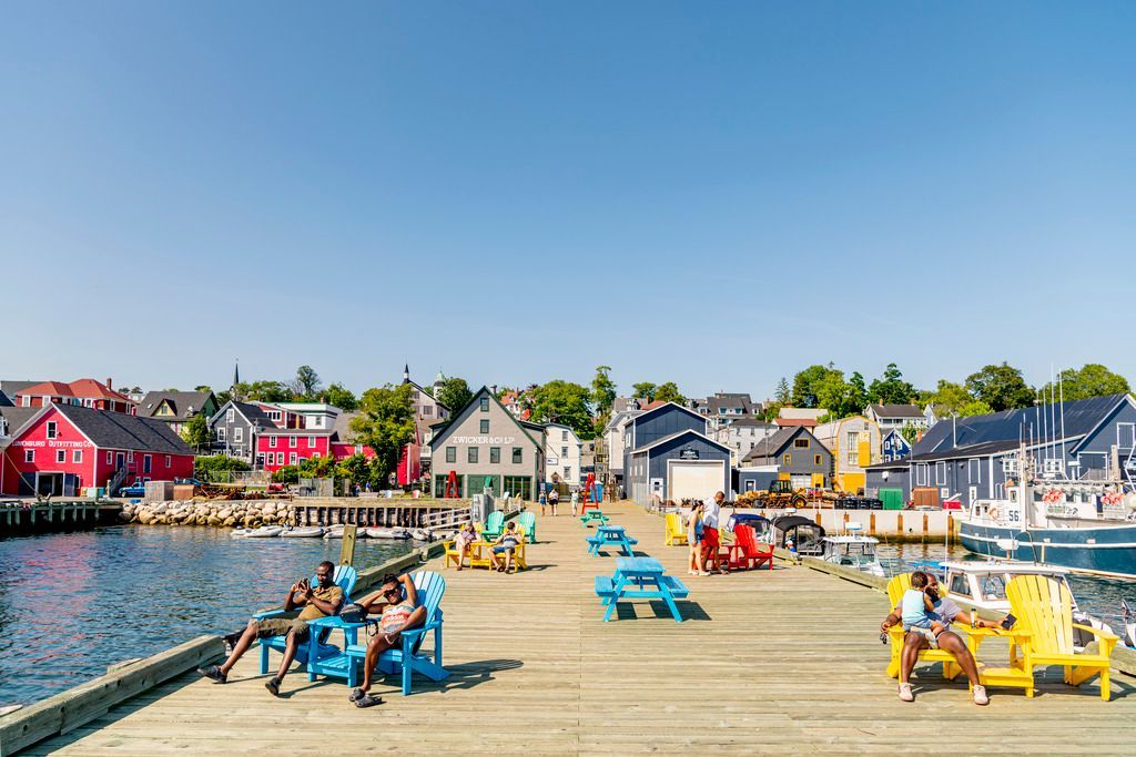 Kleurrijke ligstoelen op een zonnige houten pier aan kalm water, met huizen en boten langs de kustlijn van Lunenburg, Nova Scotia.