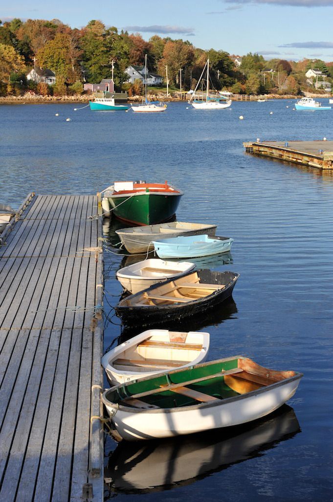 Kleine roeibootjes liggen aangemeerd aan een steiger in Mahone Bay op Nova Scotia op een kalme blauwe zee, met bomen en zeilboten op de achtergrond.