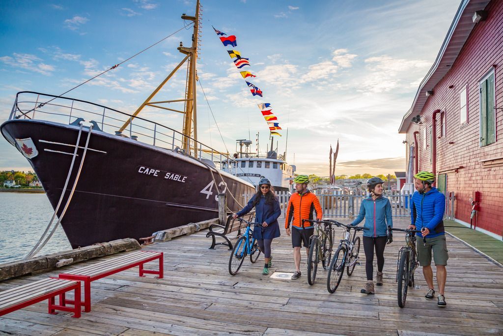 Mensen met fietsen lopen naast een groot zwart schip aan de pier van Lunenburg op Nova Scotia, met kleurrijke vlaggen erboven.