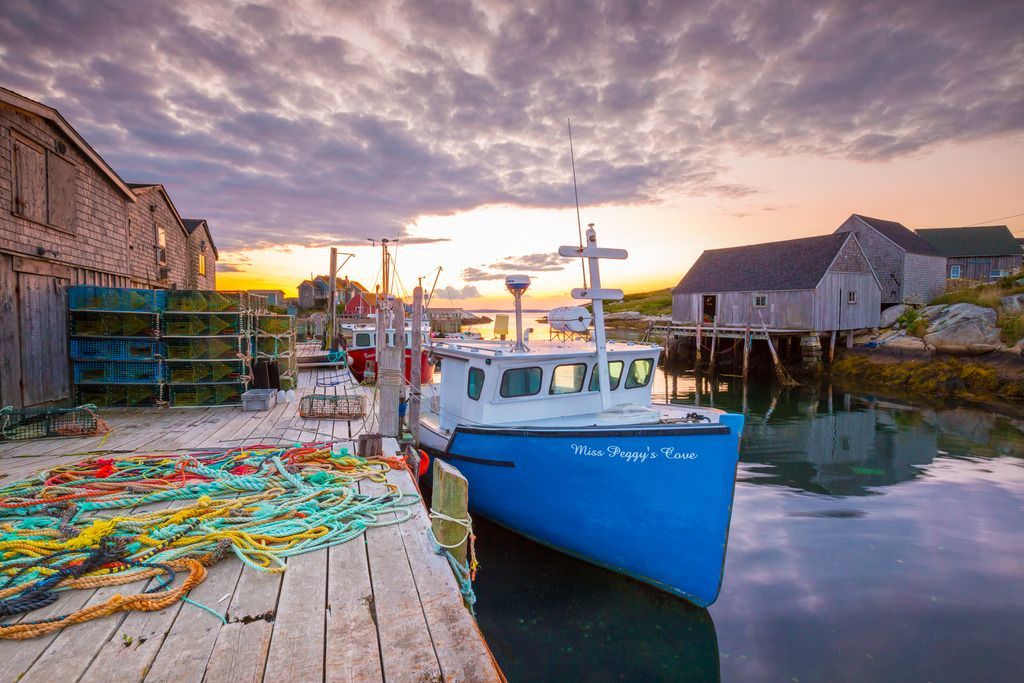 Een blauwe vissersboot ligt aangemeerd aan een steiger in Peggy's Cove op Nova Scotia bij zonsondergang, met kleurrijke netten en havengebouwen die weerspiegeld worden in het kalme water.